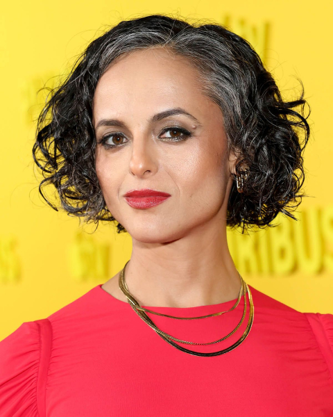 Woman with gray curly bob haircut wearing bright coral top and gold necklaces