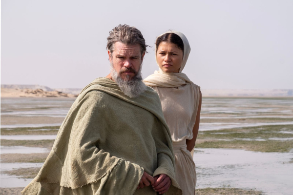 Matt Damon is Odysseus and Zendaya is Athena in THE ODYSSEY walking on a beach.